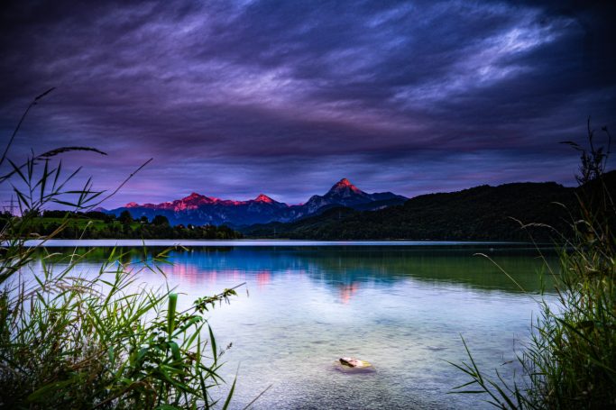 Ruhiger See mit bergiger Kulisse und Abendhimmel in lila-rot Tönen.