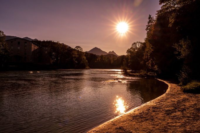 Sonnenuntergang über einem ruhigen Fluss, umgeben von Bäumen und Bergen.